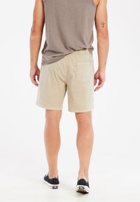 Beige katoenen shorts met een ontspannen pasvorm, elastische tailleband en een achterzak. Gecombineerd met een grijs mouwloos shirt en zwarte schoenen.