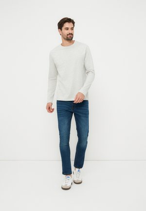 Mann trägt hellgraues Langarmshirt, blaue Jeans und weiße Turnschuhe, steht vor schlichtem weißem Hintergrund.