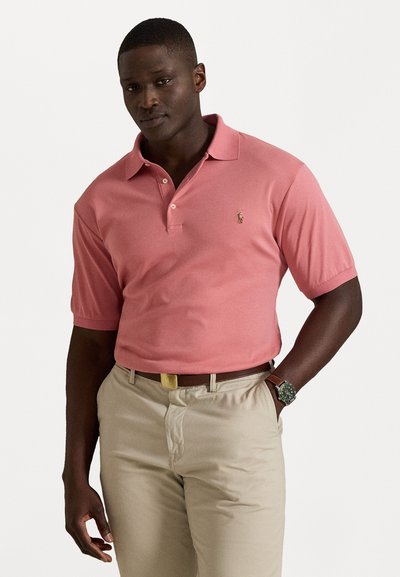 Polo de algodón en color rosa coral, de mangas cortas, con tapeta de tres botones y un pequeño logo bordado. Pantalones beige con cinturón.