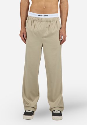 Beige wide-leg pants with elastic waistband featuring black "PEGADOR" text. Smooth texture, full-length design, paired with black sneakers.