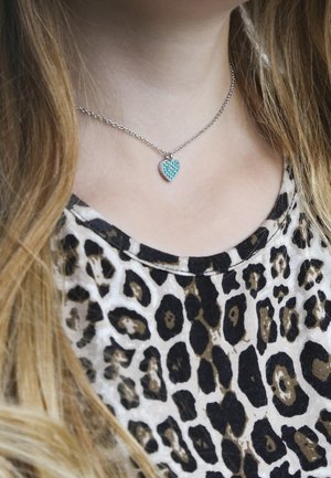 Collier en argent avec un pendentif en forme de cœur en turquoise orné de petits cristaux, porté sur une chemise à imprimé léopard.