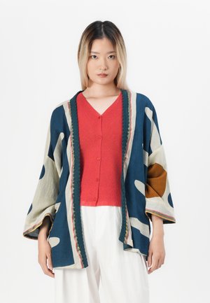 Una giovane donna con capelli lisci indossa un top rosso con bottoni, pantaloni bianchi larghi e una giacca kimono a maniche larghe con un motivo blu, beige e marrone.