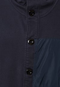 Tmavě modrá košile s texturovanou tkaninou a hladkým panelem. Dva knoflíky s lesklým povrchem na límci, kontrastní detail z látky.