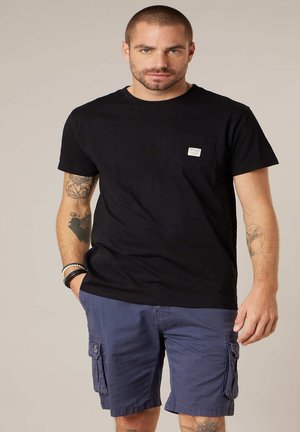 Man met kort haar en tatoeages, gekleed in een zwart T-shirt met zakje en blauwe cargobroek, staand met één hand in zijn zak.