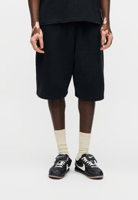 Μαύρα βαμβακερά σορτς με άνετη εφαρμογή, με απλό σχέδιο και μήκος μέχρι το γόνατο. Φορεμένα με μαύρα αθλητικά παπούτσια και μπεζ κάλτσες ψηλού τύπου.