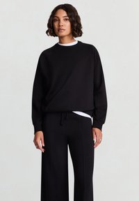 Schwarzer Pullover mit Rundhalsausschnitt und Raglanärmeln, kombiniert mit lockeren schwarzen Hosen. Mit weißer innerer Lage, die hervorblitzt.