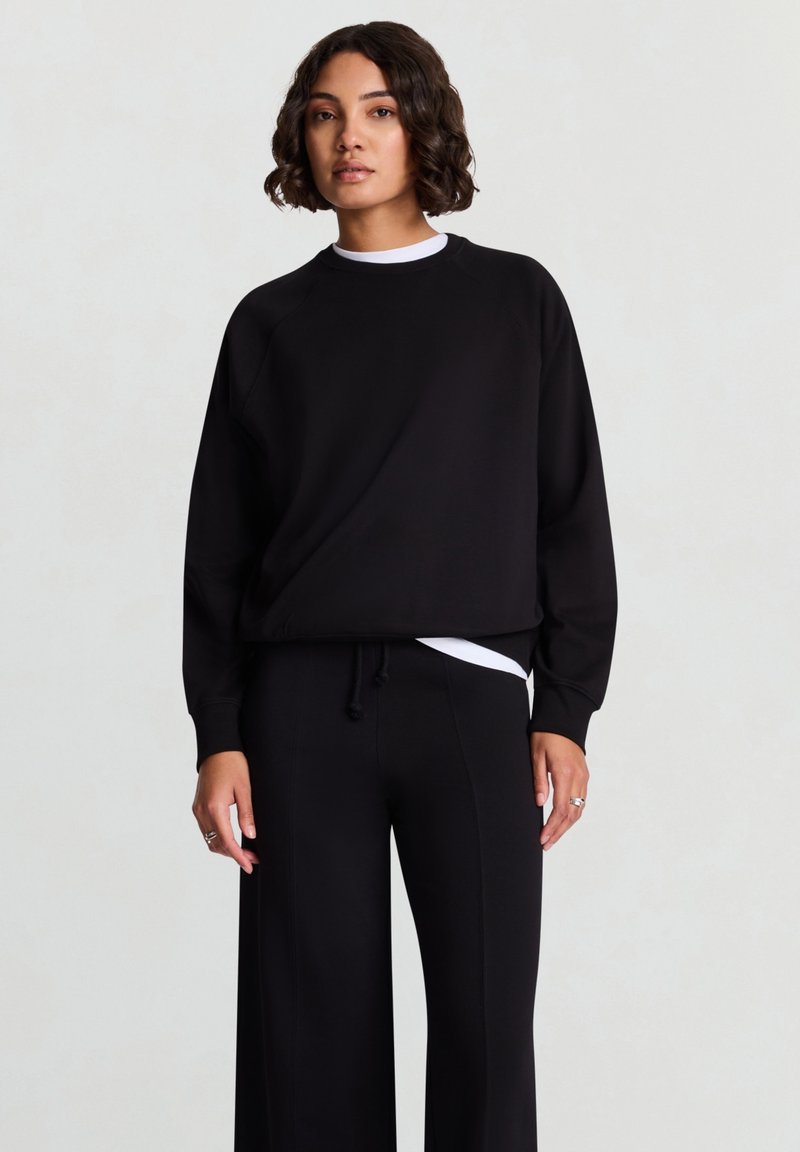 Schwarzer Pullover mit Rundhalsausschnitt und Raglanärmeln, kombiniert mit lockeren schwarzen Hosen. Mit weißer innerer Lage, die hervorblitzt.