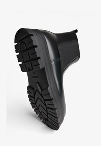 Czarny botek za kostkę z gumową podeszwą, teksturowaną powierzchnią, elastycznymi panelami po bokach i zaokrąglonym noskiem. Posiada grubą, wzorzystą bieżnikowaną podeszwę zapewniającą przyczepność.