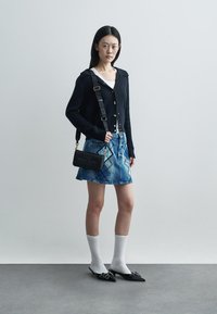 Crni rebrasti kardigan s dugmadi, bijela majica na bretele, plava traper suknja, crna mini torbica, bijele čarape do koljena i crne cipele s otvorenom petom.