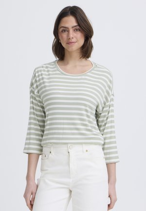 Młoda kobieta ubrana w jasnozieloną i białą koszulę w paski z długim rękawem oraz białe spodnie z wysokim stanem, stojąca na jednolitym tle.