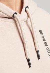 Beige hoodie heeft trekkoorden met donkere plastic aglets en een gestructureerde stof. Aan de voorkant zit een zwart gedrukte grafische afbeelding.