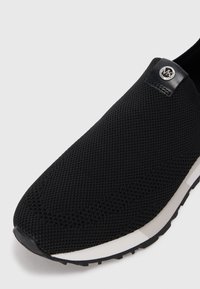 Sapatos deslizantes pretos feitos de malha respirável, com uma superfície texturizada, um detalhe em couro preto com um logótipo, e uma sola branca com sola preta antiderrapante.