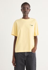 T-shirt en coton jaune à manches courtes, col rond, avec un petit logo brodé vert sur la poitrine gauche. Porté avec un jean gris foncé.