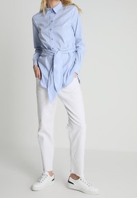 Camisa azul claro con botones en la parte delantera y detalle de cinturón, combinada con pantalones de mezclilla blancos y zapatillas blancas con un acento negro.