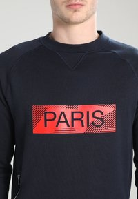 Felpa blu navy con un grande grafico rettangolare rosso che presenta la parola "PARIS" in carattere nero, con motivi testurizzati sullo sfondo rosso.