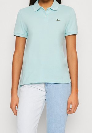 Vrouw draagt een lichtblauw poloshirt met korte mouwen en een klein groen krokodillenlogo, gecombineerd met two-tone spijkerbroeken in licht- en middelformaat blauw.