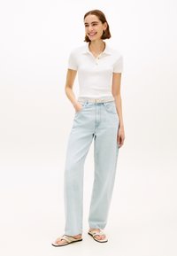 Polo blanc avec trois boutons dorés, associé à un jean large bleu clair et une ceinture blanche. Le mannequin porte des tongs beiges.