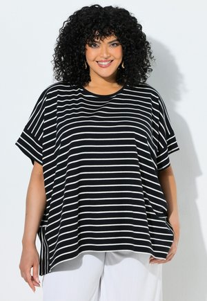 Smilende kvinde med krøllet hår, iført guld hoop-øreringe, en løs sort-hvid stribet top og hvide bukser mod en ensfarvet baggrund.