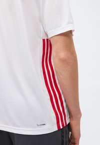 Λευκό αθλητικό πουκάμισο με κοντά μανίκια και τρεις κάθετες κόκκινες ρίγες στο πλάι, φορεμένο από άτομο με ορατό γυμνό χέρι και σκουρόχρωμο παντελόνι.