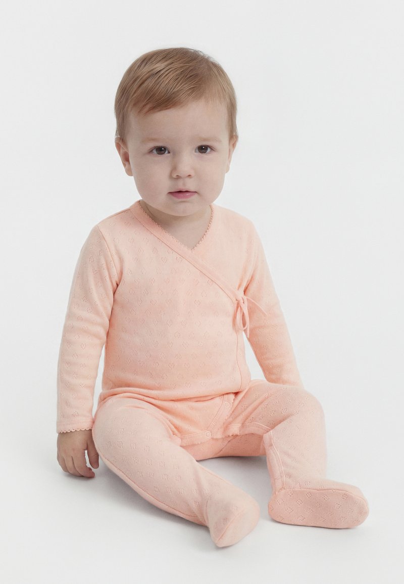Νήπιο με ανοιχτό καστανά μαλλιά καθιστό, φοράει ανοιχτό ροζ ολόσωμο φορμάκι με μακριά μανίκια και ποδαράκια, με έναν μικρό φιόγκο στο πλάι.