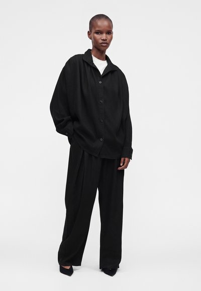 Femme portant une chemise noire ample boutonnée et un pantalon noir large avec des chaussures noires pointues sur un fond clair uni.