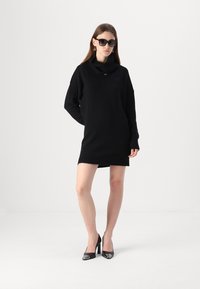 Crna pletena haljina s visokim preklopom i dugim rukavima. Model nosi crne sunčane naočale i cipele na visoku petu. Glatka tekstura, minimalistički stil.