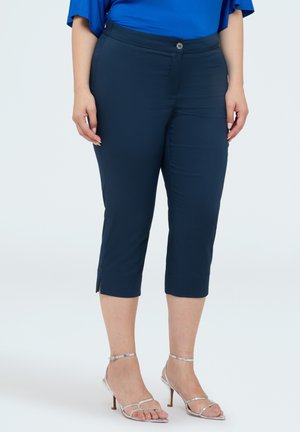 Vrouw die marineblauwe cropped broek en zilveren sandalen met hoge hakken draagt, staand tegen een effen achtergrond.