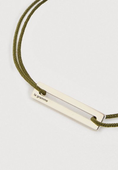 Srebrni pravokutni privjesak s ugraviranim "le gramme" na ogrlici od maslinasto zelene pletene niti na bijeloj pozadini.
