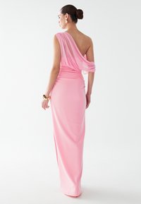 Dlouhé růžové šaty elegantně splývají, mají asymetrický design s jedním ramínkem a splývající zadní látku, s jemnou texturou a přiléhavým siluetou.