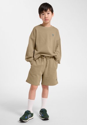 Garçon debout, les mains dans les poches, portant un sweat-shirt beige, un short assorti jusqu'aux genoux, des chaussettes blanches et des baskets vertes sur un fond blanc.