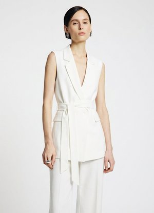 Donna con capelli corti e scuri che indossa un blazer bianco senza maniche con cintura e pantaloni bianchi coordinati su sfondo neutro.