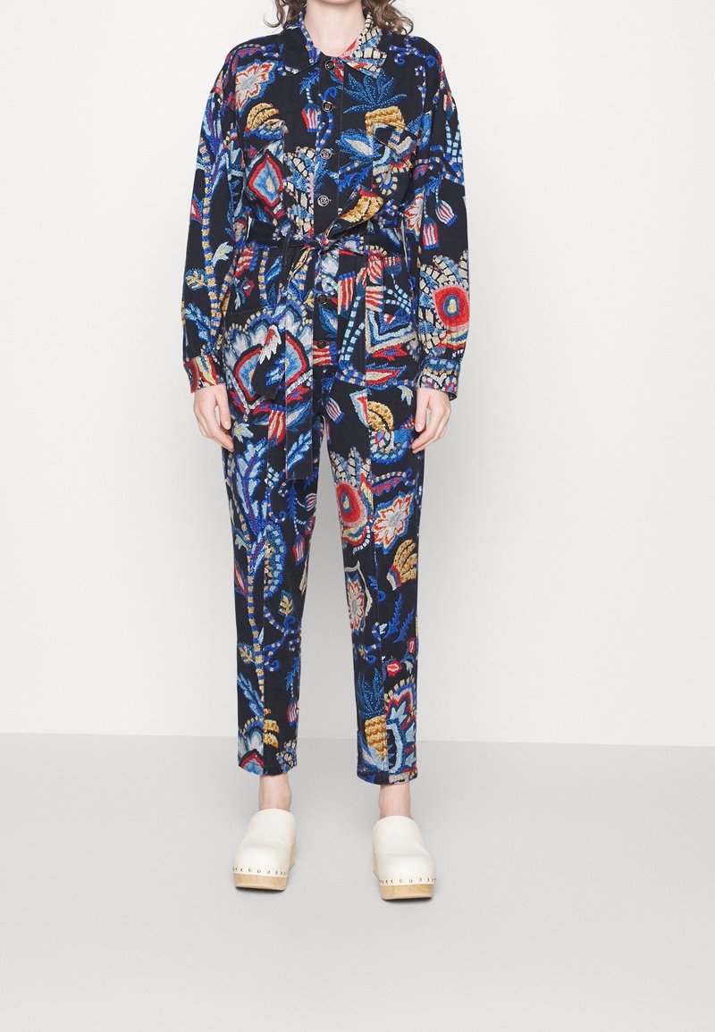 Personne portant une chemise à manches longues boutonnée assortie à un pantalon, tous deux ornés d'un motif floral abstrait et coloré, assortis à des sabots crème.