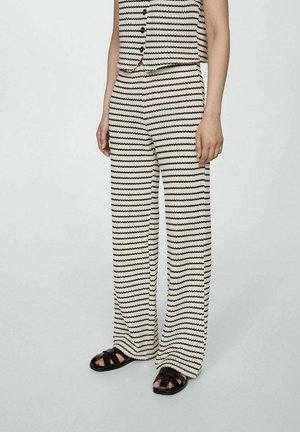Femme portant un pantalon en maille rayé noir et blanc et un gilet assorti avec des sandales noires, debout sur un sol gris clair.