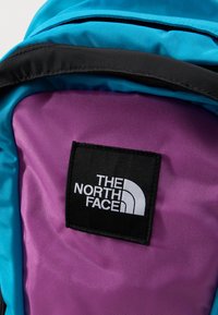 Κοντινή λήψη της μπροστινής τσέπης ενός μπλε και μοβ σακιδίου πλάτης με ένα μαύρο υφασμάτινο ταμπελάκι που δείχνει το λογότυπο The North Face σε λευκό.