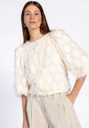 Witte textured blouse met pluizige accenten, ronde halslijn en pofmouwen. Voorzien van knopen aan de voorkant en een losse pasvorm, gecombineerd met lichtbeige broek.