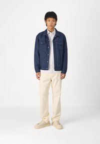 Giacca trapuntata blu marina con bottoni a pressione e due tasche sul petto, sovrapposta a una camicia bianca a righe e pantaloni color crema con sneakers beige.