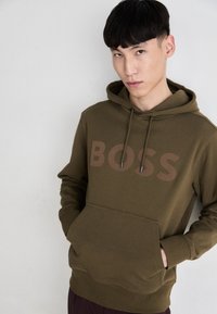 Olivegrüner Hoodie aus Baumwollmischung, mit einer Vordertasche und dem Wort "BOSS" in einer dunkleren Farbe über der Brust, mit Kordelzügen.