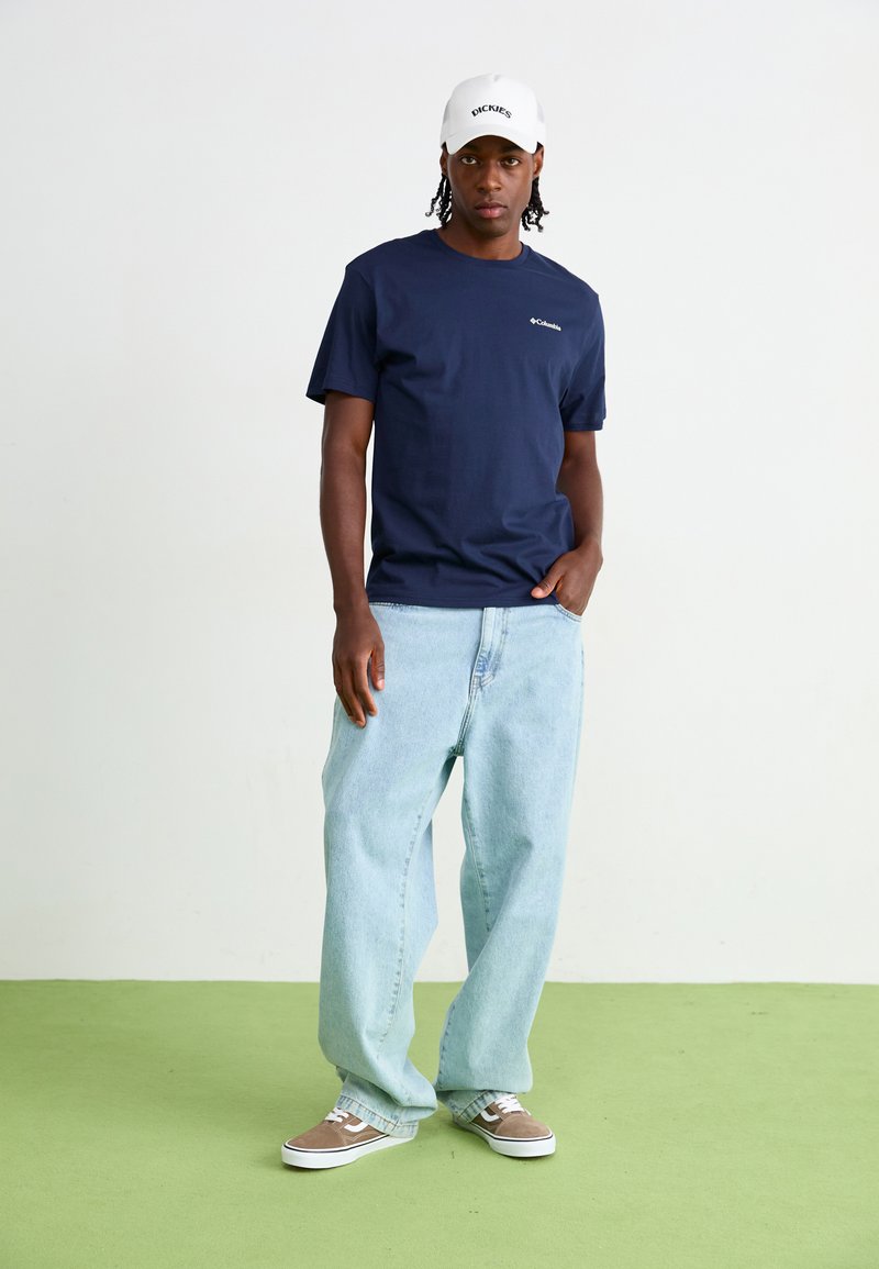 Marineblauwe katoenen t-shirt met klein logo, lichtblauwe baggy spijkerbroek en bruine sneakers met witte accenten. Witte pet met tekst. Groene achtergrond.