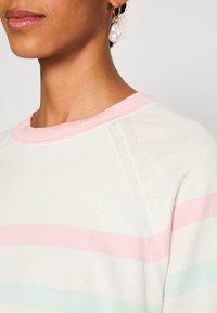 Pull crème avec des rayures horizontales rose et bleu clair, doté d'un col côtelé et d'une texture douce. Boucles d'oreilles en or avec une perle.