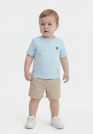 Tout-petit debout portant une chemise à manches courtes bleu clair avec un logo jaune, un short beige et des baskets blanches sur un fond uni.