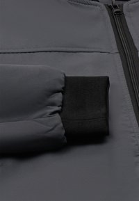 Szürke párnázott dzseki részletei, fekete bordázott mandzsettával és fekete cipzárral az elején, sima felülettel.