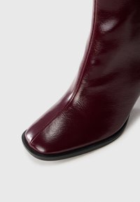 Primo piano di un boot in pelle rosso scuro con punta quadrata e suola nera su uno sfondo bianco.