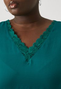 Blouse teal avec un décolleté en V orné d'une délicate bordure en dentelle festonnée. Tissu au toucher lisse avec un léger éclat. Pas de motifs visibles.