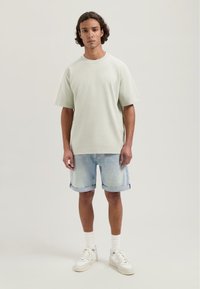 Tricou oversized verde deschis cu guler rotund, asociat cu șorturi din denim rulate și adidași albi, completat cu ciorapi albi până la gleznă.