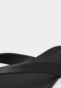 Μαύρο δερμάτινο σανδάλι με φαρδιές λουρίδες που διασταυρώνονται πάνω στο πόδι, εμφανιζόμενο σε κοντινό πλάνο πάνω σε απλό ανοιχτό φόντο.