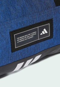Blue textured fabric bag with a black label featuring white text and three white stripes at the bottom. Sports brand logo visible.