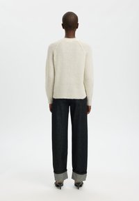 Krém színű kötött pulóver raglán ujjakkal, sötétkék széles szárú farmerral, hajtott szegéllyel és hegyes orrú sarkú cipővel párosítva.