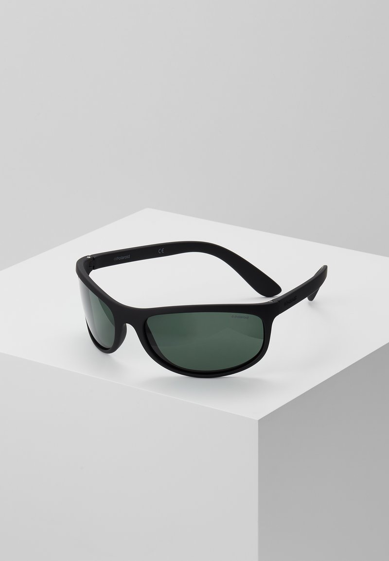 Lunettes de soleil noires avec des verres teintés en vert. Fabriquées en plastique, elles présentent un design épuré et une monture courbée pour un maintien sécurisé. Marquées CE.