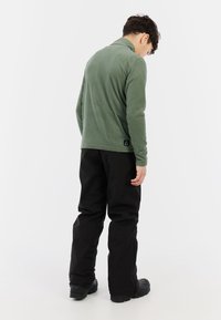 Pullover verde in pile con colletto alto e pantaloni neri. Il tessuto sembra morbido, con una texture liscia e minimi dettagli di cucitura. Calzature nere.