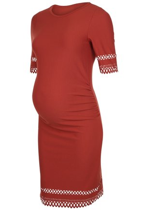 Zwangere vrouw in een nauwsluitende roestrode jurk met kanten afwerking, draagt een zwarte jas over haar schouder, en zwarte schoenen met lage hak.